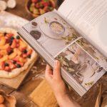 A person reads a cookbook while preparing a fruit tart on a wooden table.